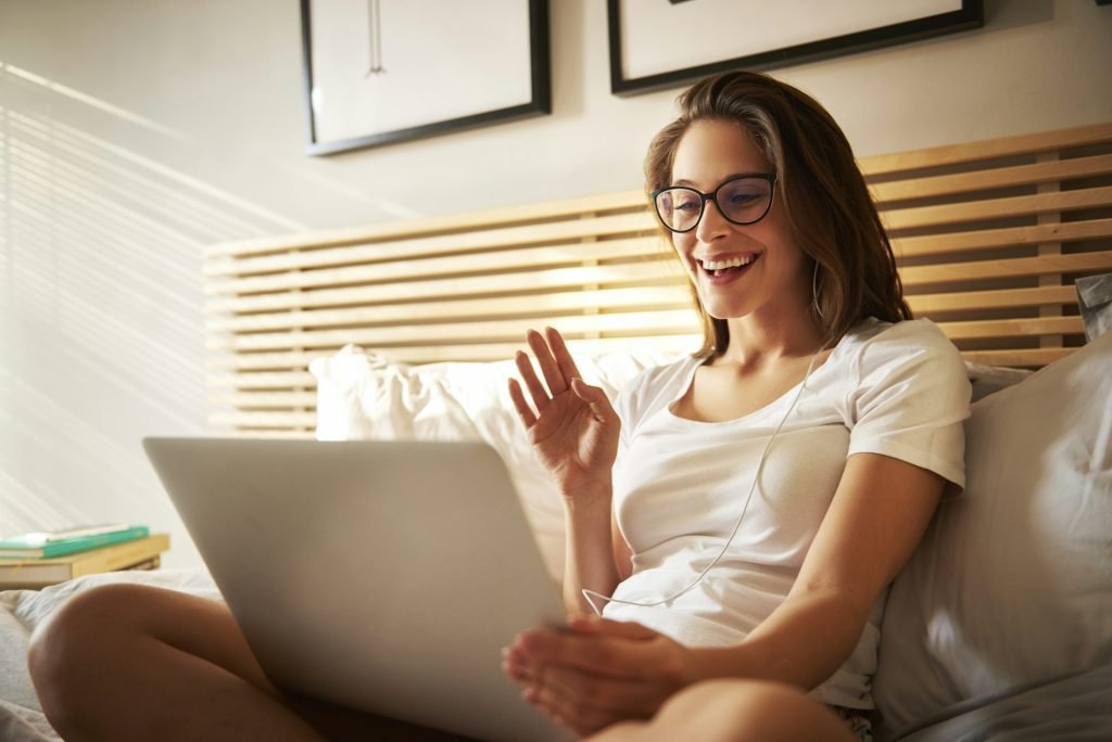Portrait of laughing young woman sitting on bed with laptop chatting